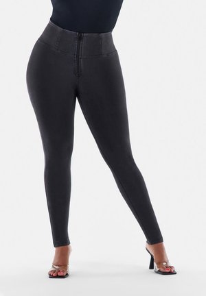 Nainen, jolla on korkeavyötäröiset mustat kapeat farkut, joissa on etuvetoketju, ja kirkasremmillä varustetut korkokengät, seisoo jalat ristissä.