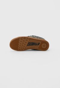 Podošva čierneho a hnedého tenisky ležiaca na boku, s vyrazeným nápisom "ETNIES" a kruhovým logom na gumovej podrážke.