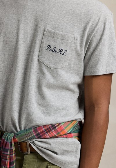 Close-up of person wearing a light gray T-shirt with "Polo RL" embroidered on the chest pocket and a colorful plaid shirt tied around the waist.
