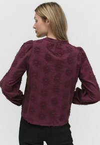 Bordeaux blouse met bloemenborduursels, pofmouwen en een ronde halslijn. Geweven stof met ingewikkelde details langs de achterkant.