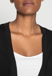 Schwarzer Cardigan über einem weißen Top mit V-Ausschnitt. Glatte Textur, offene Vorderseite und lange Ärmel, die vom Halsausschnitt abwärts sichtbar sind.