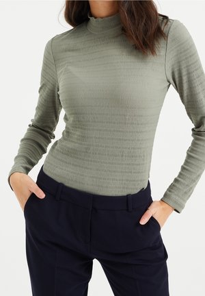 Kobieta w teksturowanej oliwkowozielonej bluzce z długim rękawem wpuszczonej w czarne, dopasowane spodnie, z rękami w kieszeniach, na białym tle.