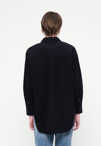 Cămașă din corduroy bleumarin cu mâneci lungi, guler ascuțit și manșete cu nasturi. Are o croială dreptă și o cusătură centrală pe spate.
