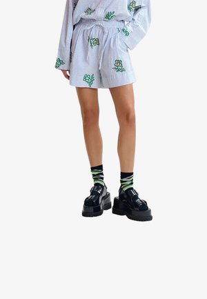 Pantaloncini a righe e maglia a maniche lunghe con stampa floreale verde, abbinati a sandali platform neri con suola spessa e calzini con motivo nero, verde e bianco.