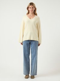 Pull crème à col en V avec des manches longues associé à un jean large bleu clair. La tenue est complétée par des chaussures en daim marron.