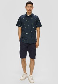 Korte mouwen marineblauw shirt met groene palmboomprint, knoopsluiting aan de voorkant, gecombineerd met donkere cargoshorts en lichtbeige sneakers.