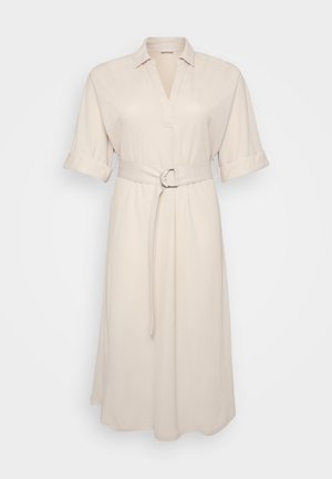 Bežs krekla kleita ar apkakli un sarullētām piedurknēm, ar jostas siksnu ar apaļu sprādzi, karājas uz vienkārša fona.