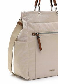 Beige Tragetasche mit vordere Reißverschlusstasche, braunem Leder-Reißverschlussanhänger, Seitentasche und zwei braunen Ledergriffen mit silberner Beschlagsarbeit.