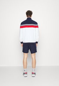 Tracksuit jacket in light gray with navy accents and red stripe, paired with navy shorts; fabric appears smooth with ribbed cuffs.