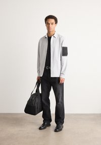Cămașă butonată de un gri deschis, cu un buzunar negru pe brațul stâng, tricou negru dedesubt, blugi negri și pantofi negri. Ține o geantă neagră.