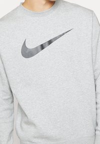 Grå sweatshirt gjord av fleece-material, med en stor svart Nike swoosh-logotyp. Enkel design med rund hals och ribbade ärmslut och fåll.