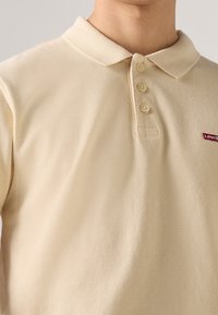 Polo beige à manches courtes avec un tissage texturé, trois boutons et un petit logo rouge Levi's sur la poitrine gauche. Design de col classique.