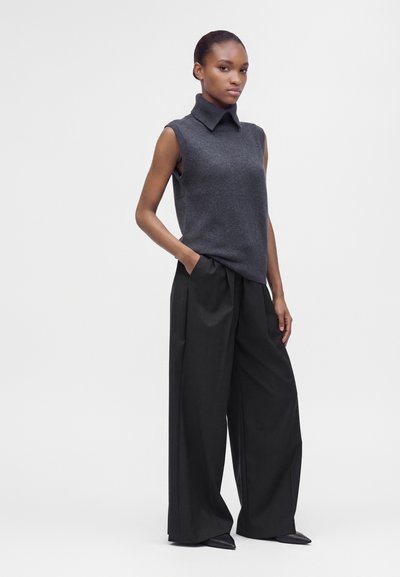 Femme portant un haut en maille gris foncé sans manches avec un large col et un pantalon noir à jambes larges, debout avec une main dans la poche.