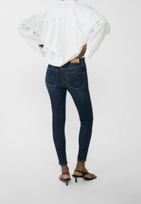 Persona vestida con jeans ajustados azul oscuro, una camisa de manga larga blanca y sandalias negras de tacón, de pie con las manos en las caderas contra un fondo blanco.