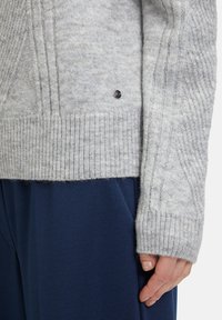 Hellgrauer Strickpullover mit gerippten Bündchen und Saum, strukturiertem Muster und kleinen dunklen Knopfdetails, kombiniert mit marineblauen Hosen.