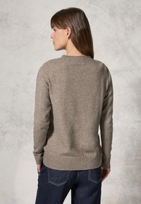 Hellbraunes gestricktes Sweatshirt mit ribbelndem Rundhalsausschnitt und Bündchen, vor einem strukturierten Hintergrund getragen, kombiniert mit dunklen Jeans.
