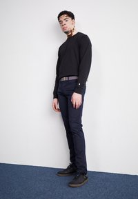 Chemise noire à manches longues avec une coupe ajustée, portée avec un pantalon ajusté bleu marine et des baskets noires. Comprend une boucle de ceinture rectangulaire en argent.