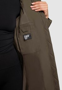 Chaqueta acolchada en verde oliva con un bolsillo interior visible, que presenta una etiqueta negra con texto blanco, hecha de un material suave y ligero.