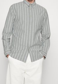Jasnoszara koszula z długim rękawem i pionowymi białymi paskami, z pojedynczą kieszenią na piersi, w zestawie z białymi spodniami.