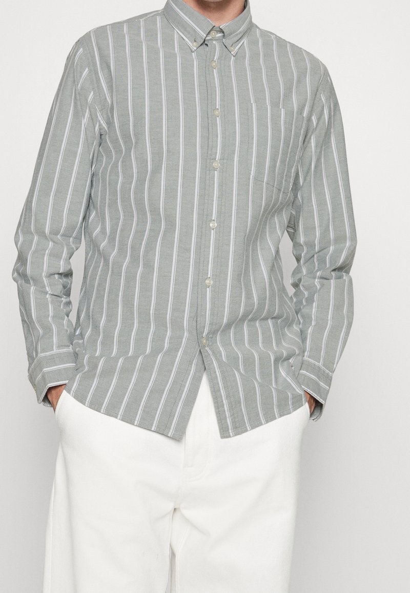 Jasnoszara koszula z długim rękawem i pionowymi białymi paskami, z pojedynczą kieszenią na piersi, w zestawie z białymi spodniami.