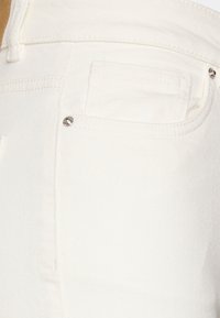 Tela de mezclilla blanca, primer plano de un bolsillo trasero con un pequeño adorno de remache plateado, con costuras cosidas y una textura suave.