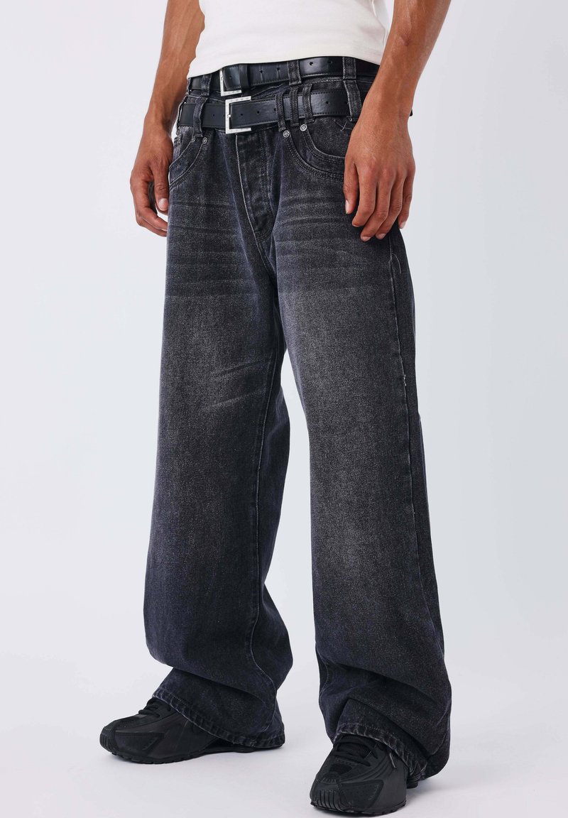 Jeans negros de pierna ancha hechos de mezclilla con una textura desgastada, con trabillas para cinturón y bolsillos delanteros, combinados con zapatillas negras.