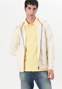 Beige hoodie met rits en oranje accenten, gecombineerd met een lichtgele polo. Casual uitstraling met een ontspannen pasvorm.