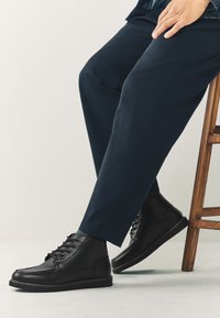 Schwarze Leder-Stiefeletten mit Schnürdesign, glatter Oberfläche und Steppdetail am Schaft. Kombiniert mit dunklen, schlank geschnittenen Hosen.