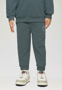 Donkergroene joggingbroek met geribbelde boorden en tailleband, voorzien van een klein wit logo op de bovenbeen, gecombineerd met witte sneakers.
