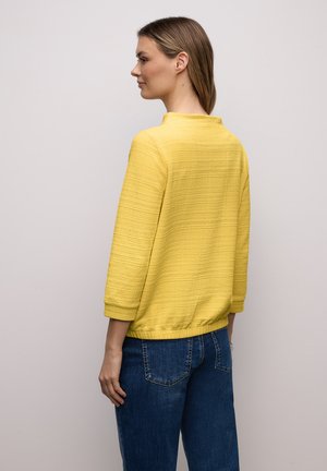 Femme vue de dos portant un pull jaune texturé à manches trois-quarts et un jean bleu sur un fond clair uni.