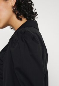 Camisa preta com colarinho, featuring mangas bufantes, tecido suave e um corte ajustado, realçando linhas elegantes e um design minimalista.