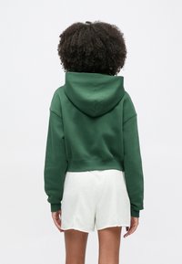 Sweat à capuche vert court en tissu doux avec capuche à cordon de serrage et ourlet côtelé, associé à un short de couleur claire. Surface texturée visible.