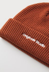 Beanie muts in roestoranje, geribbeld textuur met een omgeslagen boord. Heeft "Originals Studio" geborduurd in het wit.