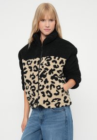 Svart og beige fleecejakke med leopardmønster på den nederste halvdelen, glidelås i front og høy krage. Har avrundede ermer og lommer på siden.