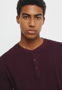 Bordowa long-sleeve henley z teksturowanej tkaniny, z okrągłym dekoltem i trzema guzikami z przodu. Zbliżenie na koszulę.