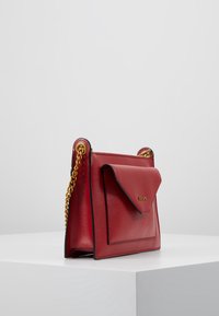 Borsa a tracolla rossa in pelle con tasca a busta frontale, tracolle in catena dorata e logo DKNY, posizionata su una superficie bianca contro uno sfondo chiaro e semplice.