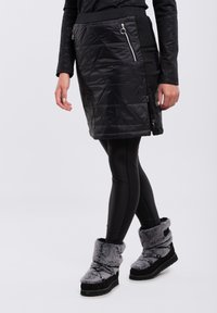 Svart quiltad kjol med sidofickor, i kombination med svarta leggings och grå plyschstövlar vid anklarna. Slät textur och åtsittande design.