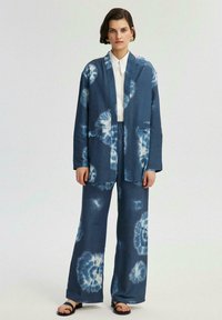 Conjunto de cárdigan y pantalones en tie-dye azul, con una textura suave, corte recto y dos bolsillos frontales. Lleva puesto una camisa blanca.