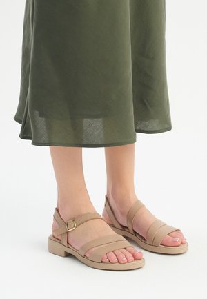 Sandali piatti beige con due cinturini larghi e dettagli a fibbia. Suola testurizzata e design aperto che mostra le dita dei piedi. Indossati con una gonna verde fluida.