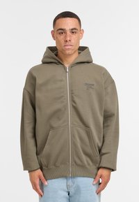 Ärmelväska i olivgrönt med dragkedja, tillverkad av mjukt material med en avslappnad passform, utrustad med en framficka, ribbade ärmslut och huva. Litet tryckt logotyp på bröstet.