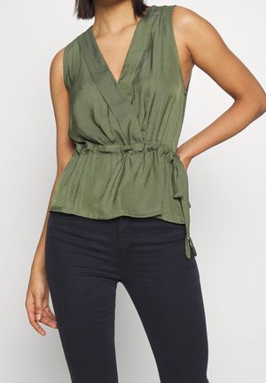 Kvinde iført ærmeløs olivengrøn slå-om top med samlet talje og sorte tætsiddende bukser, stående med den ene hånd på hoften.