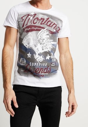 Mann trägt weißes kurzärmeliges T-Shirt mit dem Text "Montana Superior Legends" und einer Grafik einer geflügelten Trompete auf der Vorderseite, kombiniert mit schwarzen Hosen.