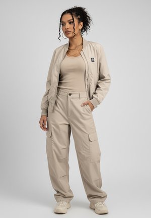 Beige bomber jacket paired with a fitted beige tank top and loose-fitting beige cargo pants, featuring pocket details and cuffed sleeves.