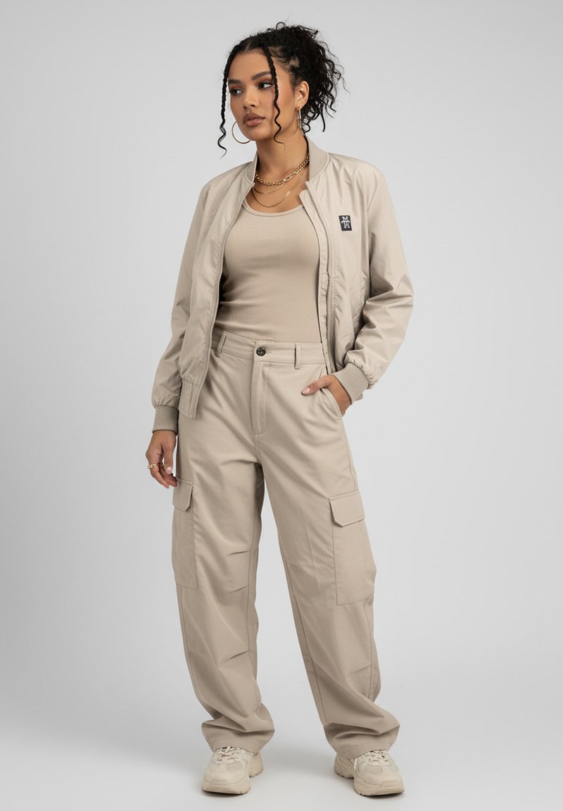 Beige Bomberjacke kombiniert mit einem enganliegenden beigen Tanktop und lockeren beigen Cargo-Hosen, die über Taschen und umschlagbare Ärmel verfügen.