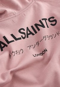 Różowy materiał z czarnym napisem "ALLSAINTS," japońskimi znakami i napisem "LONDON", ukazujący część dekoltu bluzy z kapturem.