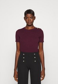 Bordeauxkleurige korte mouw gebreide top met een textuurafwerking en gouden knoopdetails op de schouder, gecombineerd met zwart, hoog getailleerde broek.