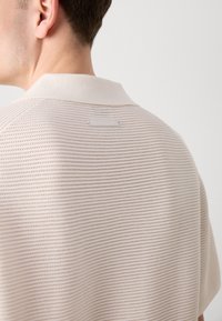 Polo-skjorte i lys beige, med et teksturert strikkedesign, korte ermer og krage; merkeetiketten er synlig bak.
