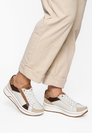 Feet wearing white, brown, and beige lace-up sneakers paired with beige, rolled-up corduroy pants on a white background.