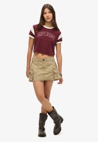 Afgekorte bordeauxrode t-shirt met witte accenten, met het "SUPERDRY" logo; kaki cargominirok met zakken; zwarte laarzen tot halverwege de kuit.