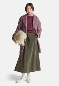 Kabát s rybí kostrou v tmavě červené barvě, fialový rolák, olivově zelená sukně a bílé tenisky s texturovanými akcenty. Modelka drží huňatý doplněk.